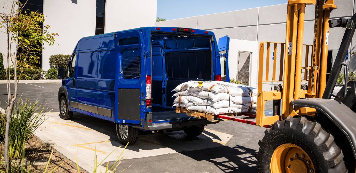 Se muestra el perfil de atrás inclinado de una Ram ProMaster 2500/3500 SLT Cargo Van High Roof 2024 azul con las puertas de atrás completamente abiertas y un montón de materiales embolsados que están cargando en el área de carga por una carretilla elevadora.