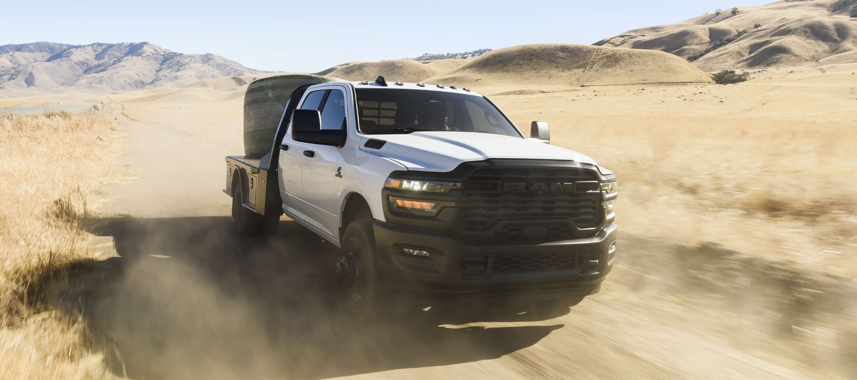 Una Ram 3500 Tradesman Chassis Crew Cab 2025 blanca cruzando el desierto con un enorme fardo de heno en su plataforma mejorada.